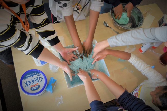 An overhead photograph of many hands reaching in to a big bowl full of light blue coloured slime.